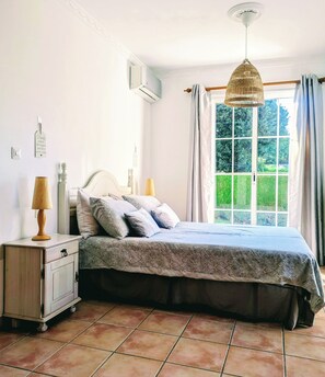A bedroom with a bed, bedside lamps, a nightstand, a window with curtains, and a view of greenery outside.