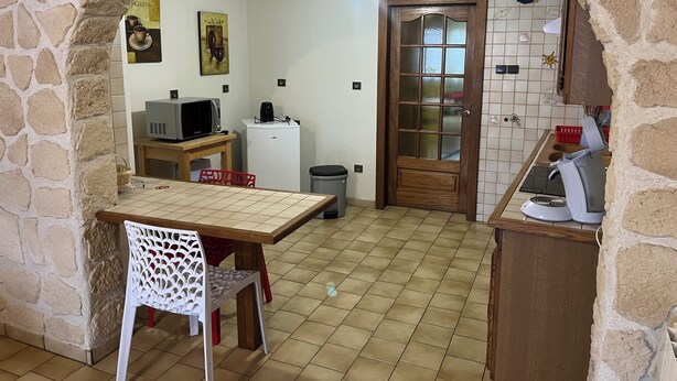 Een keuken met een stenen boog, een houten tafel, een witte stoel, een magnetron, een koelkast en een houten deur met ruitpanelen.