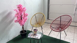 Een bloeiende roze plant in pot, twee stoelen met een draadframe en een glazen bijzettafeltje met een krant.