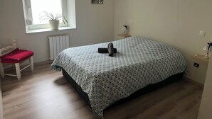 Een slaapkamer met een bed, een raam met een plant, een radiator, een stoel en een nachtkastje met een lamp.