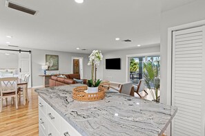 A modern kitchen with a marble countertop, wooden dining table, and chairs. There is a sofa and a TV in the living area. - Take it Keysie, Waterfront House (Marathon)