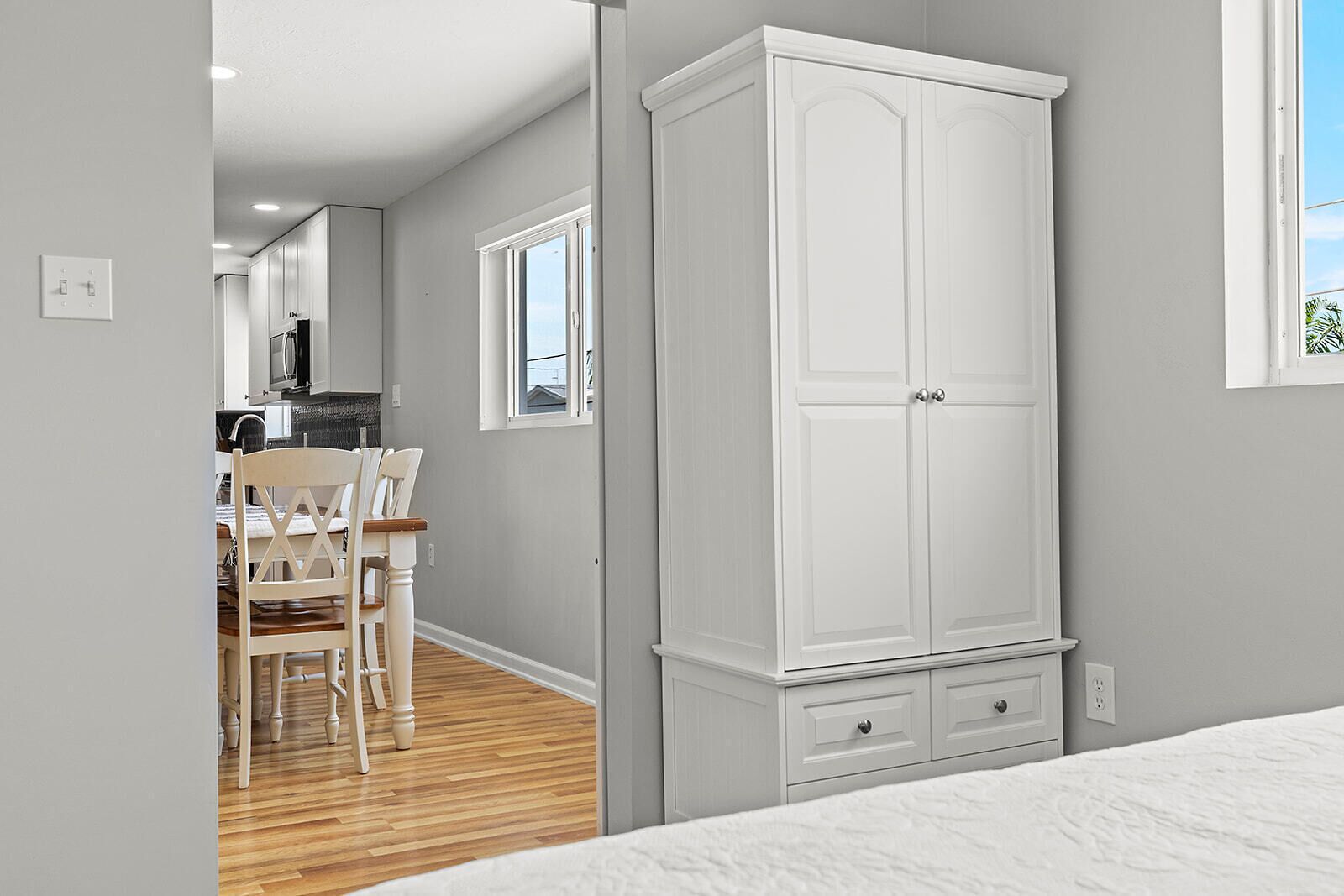 Een slaapkamer met een bed, een witte inbouwkast, een houten vloer en uitzicht op een keuken met een eettafel en stoelen.