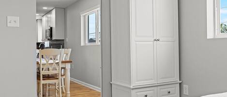 Een slaapkamer met een bed, een witte inbouwkast, een houten vloer en uitzicht op een keuken met een eettafel en stoelen.