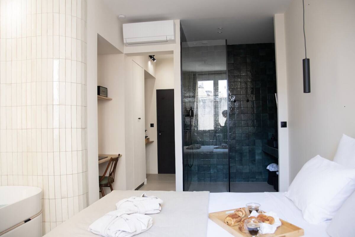Ein modernes Hotelzimmer mit einem großen Bett, einem Nachttisch mit einem Tablett mit Croissants und Kaffee, einem Badezimmer mit einer Duschkabine aus Glas und Blick auf die Stadt durch die Duschwand.