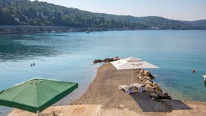 A pebble beach with two umbrellas and lounge chairs, overlooking a calm lake with boats and a forested hill in the background. - Studio Apartment 6 for two persons (Omisalj)