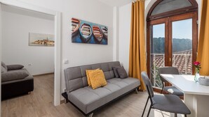 A modern living room with a grey sofa, a dining table with two chairs, a painting of boats, and a view of a cityscape through a large window with yellow curtains. - One Bedroom Apartment 5 with Sea view (Omisalj)