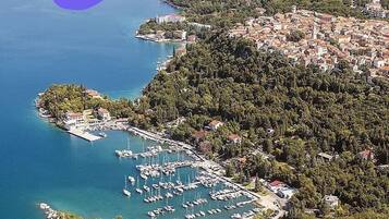 An aerial view of a coastal town with a marina, dense forested hills, and a clear blue sea.