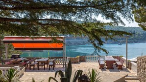 Un espace extérieur aménagé avec des tables et des chaises donnant sur un plan d’eau, un espace de repos couvert et un mur en pierre.