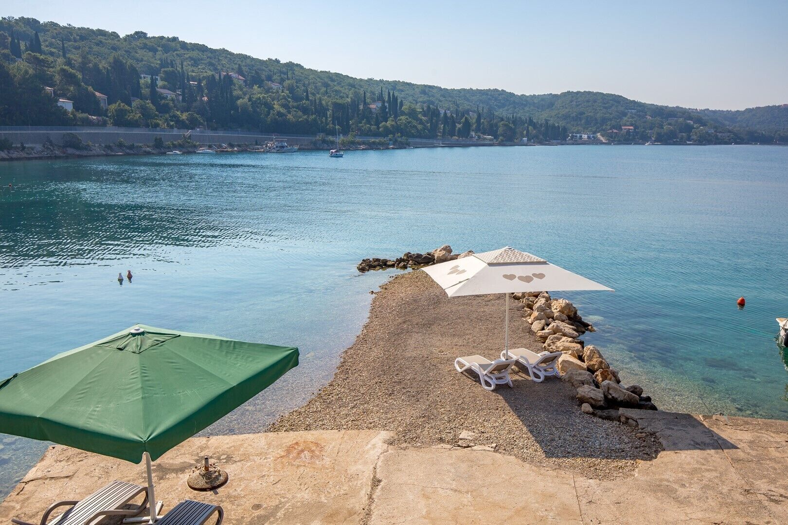 A pebble beach with two umbrellas and lounge chairs, overlooking a calm lake with boats and a forested hill in the background.