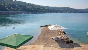 Une plage de galets avec deux parasols et des chaises longues, donnant sur un lac paisible où l’on aperçoit des bateaux, et sur une colline boisée en arrière-plan.