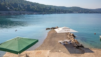 A pebble beach with two umbrellas and lounge chairs, overlooking a calm lake with boats and a forested hill in the background.