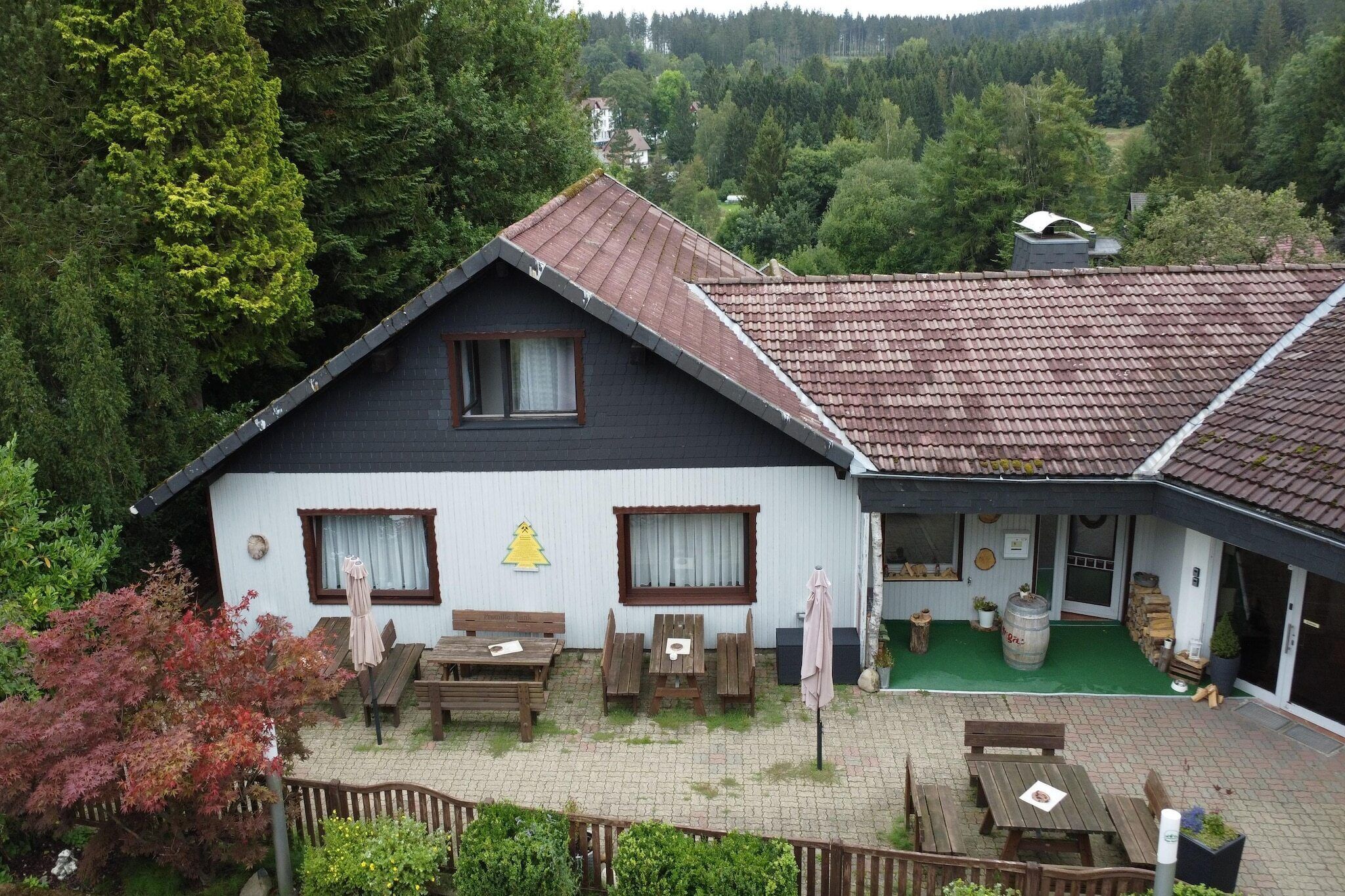A two-story chalet with a tiled roof, a patio with wooden furniture, and a view of a forested valley.