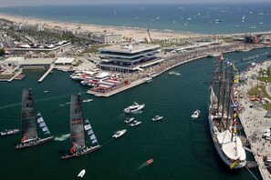 A marina with multiple sailboats, a large building with a modern design, and a beach in the background.