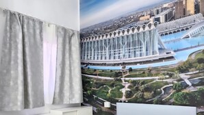 Un dormitorio con cama, ventana con cortinas, cabecero fijado a la pared y un gran mural con vista a la ciudad.