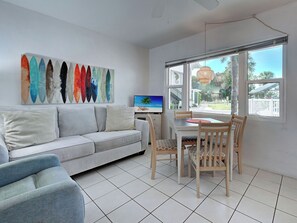 A living room with a sofa, a dining table with chairs, a TV, and a large window with a view of the outdoors.