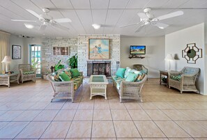 A spacious living room with wicker furniture, a fireplace, ceiling fans, and a television.