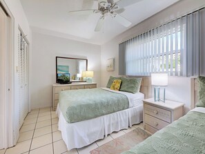A bedroom with a bed, bedside tables, a ceiling fan, a window with blinds, and a door with a shutter.