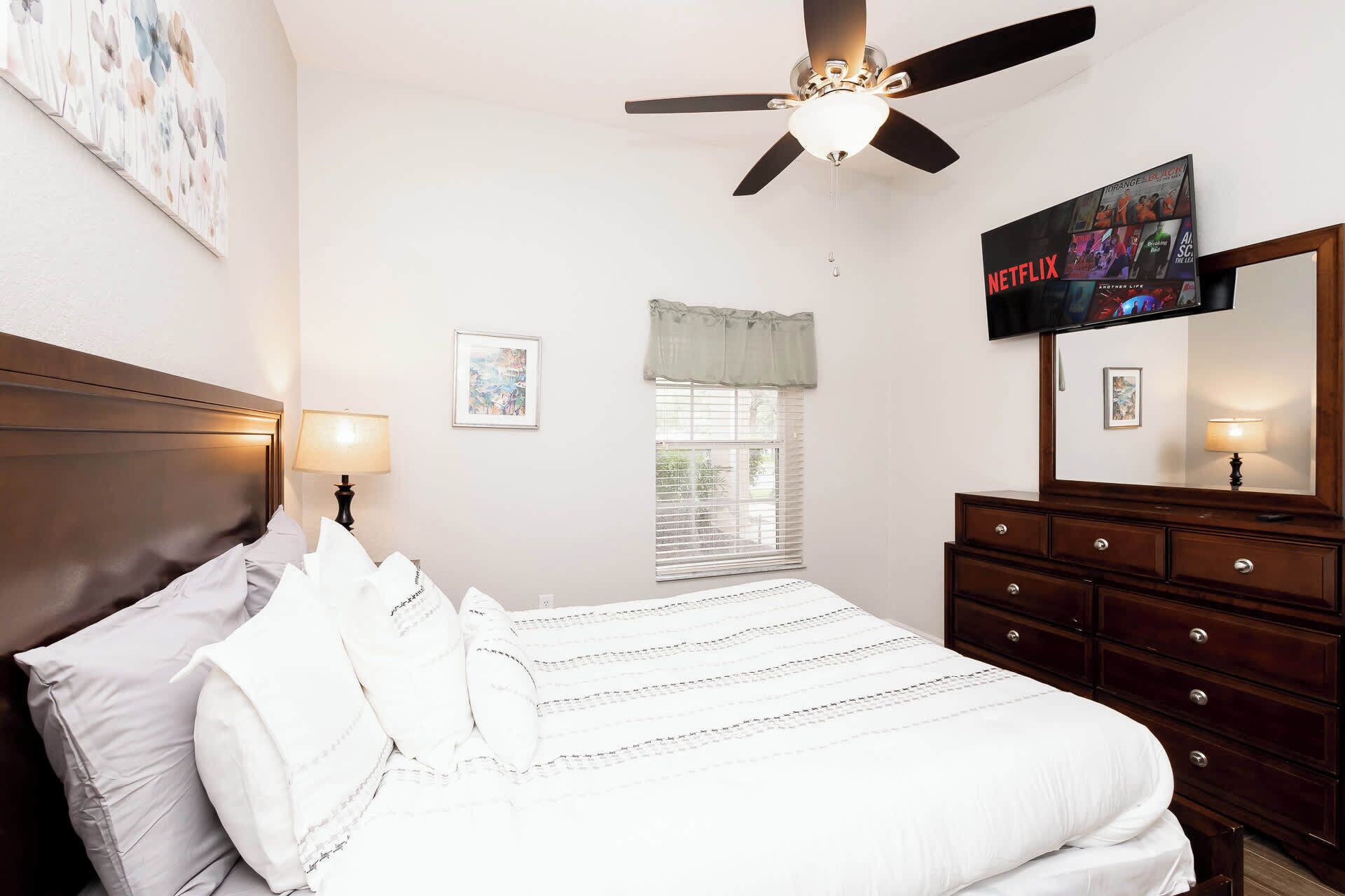 A bedroom with a bed, a dresser, a ceiling fan, a TV, and a window with blinds.