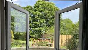 Vista attraverso porte in vetro aperte su un patio con tavolo e sedie in legno, circondato da vegetazione e una recinzione di legno.