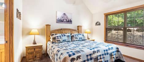Un dormitorio con una cama grande, cabecera de madera, dos mesitas de noche con lámparas, ventilador de techo, ventana con persianas y un cuadro colgado en la pared.