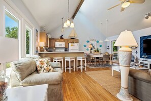 Un salon moderne avec un canapé, une table basse et un grand lampadaire. Il y a également un coin cuisine équipé d’un réfrigérateur, d’un four à micro-ondes et de meubles de rangement en bois. La pièce est dotée de grandes fenêtres et d’un ventilateur de plafond.