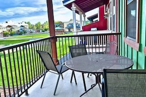 A covered outdoor seating area with a table and chairs, a railing, and a view of a green lawn and buildings in the background.