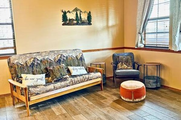 A room with a wooden floor, a sofa with patterned cushions, a chair, a round ottoman, and a wall-mounted decoration.