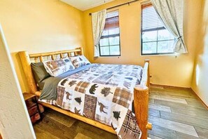 A single bed with a wooden frame and a quilted bedspread, a window with curtains, and a wooden floor.