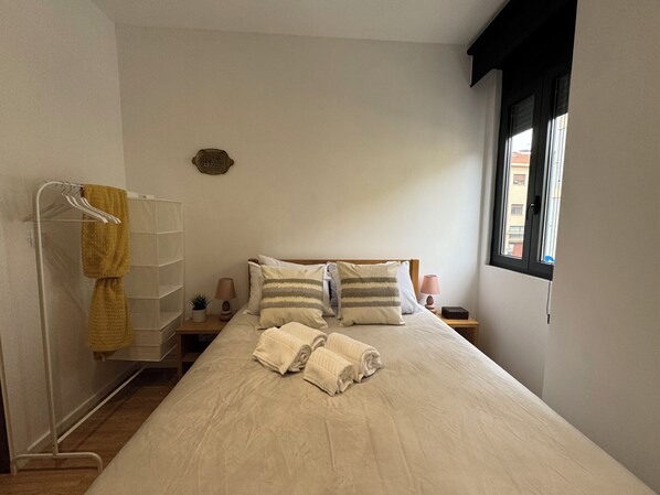 A neatly made bed with pillows and towels, a bedside table with a lamp, a wall-mounted shelf, and a window with a view of buildings. - Casas da Corujeira 2 (Porto)