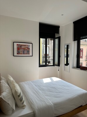 Un dormitorio con cama, almohadas, un cuadro enmarco y ventanas con contraventanas negras.