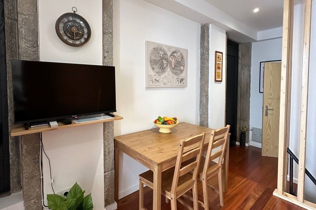 Een eetruimte met een houten tafel, stoelen, een fruitschaal, een hangklok en een flatscreen televisie.