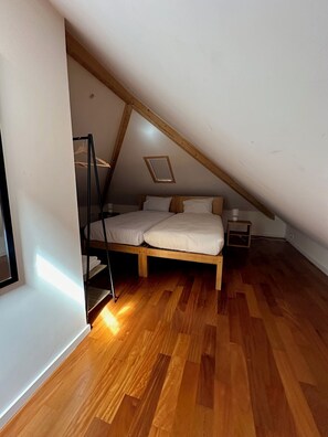 Un dormitorio con techo inclinado, suelo de madera, una cama con ropa de cama blanca, una mesita de noche pequeña y una lámpara colgante.