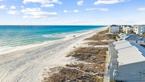 A beachside view with a sandy beach, ocean, and a row of white buildings with blue roofs. - Relax in Style Steps to the Beach • Heated Pool, Gulf Views, & Room for Everyone (Inlet Beach)