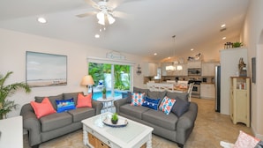 A spacious living room with a grey sofa set, a coffee table, and a ceiling fan. A kitchen is visible in the background with white cabinets and a dining area. - Family Home with Private Pool, 3 mins from Beach! - Ariel's by the Sea (Holmes Beach)