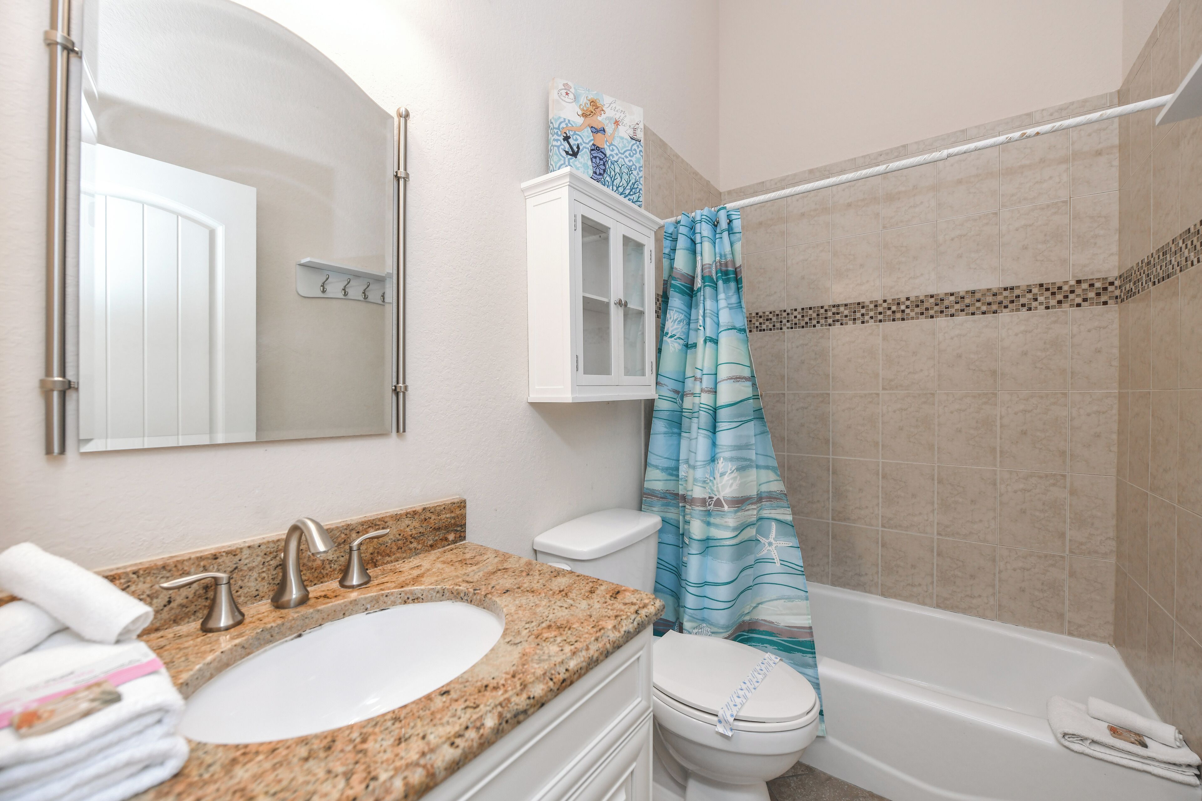 A bathroom with a granite countertop sink, a white toilet, a white cabinet, a shower curtain with a sea theme, and a bathtub.