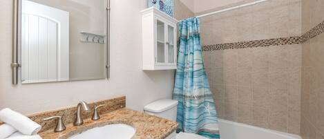 A bathroom with a granite countertop sink, a white toilet, a white cabinet, a shower curtain with a sea theme, and a bathtub.