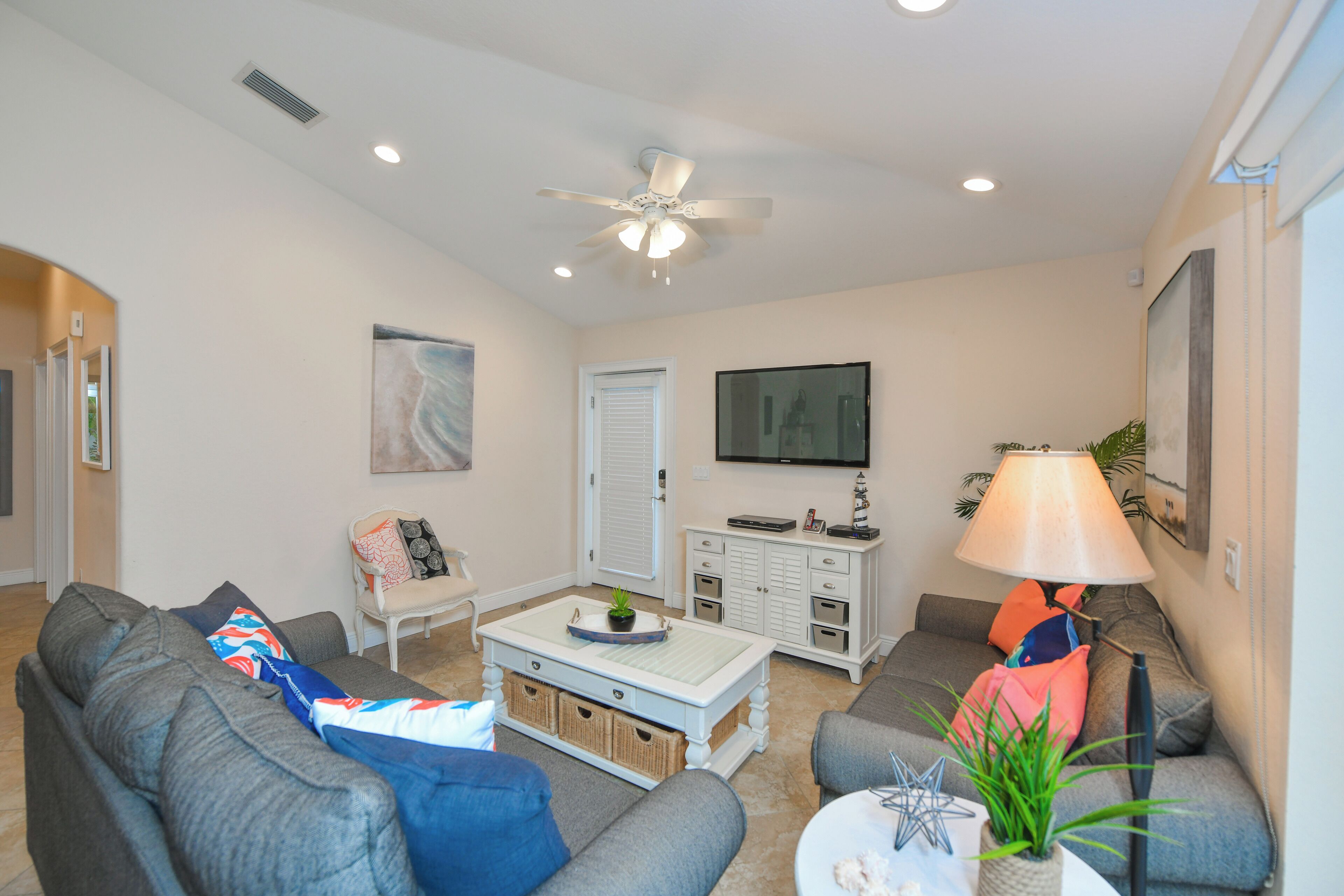 A living room with a ceiling fan, a television mounted on the wall, a coffee table with decorative items, and a sofa with cushions.