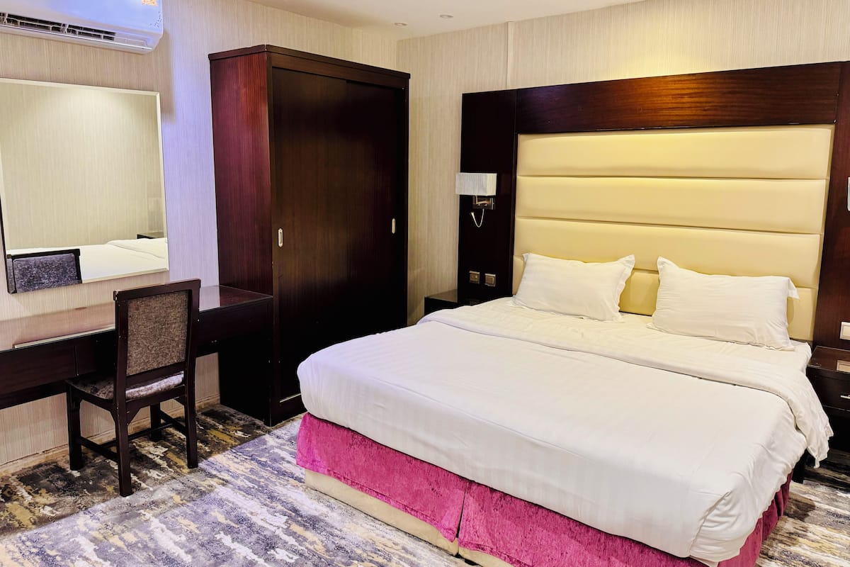 A hotel room with a large bed, a desk, a chair, a wardrobe, and an air conditioning unit.