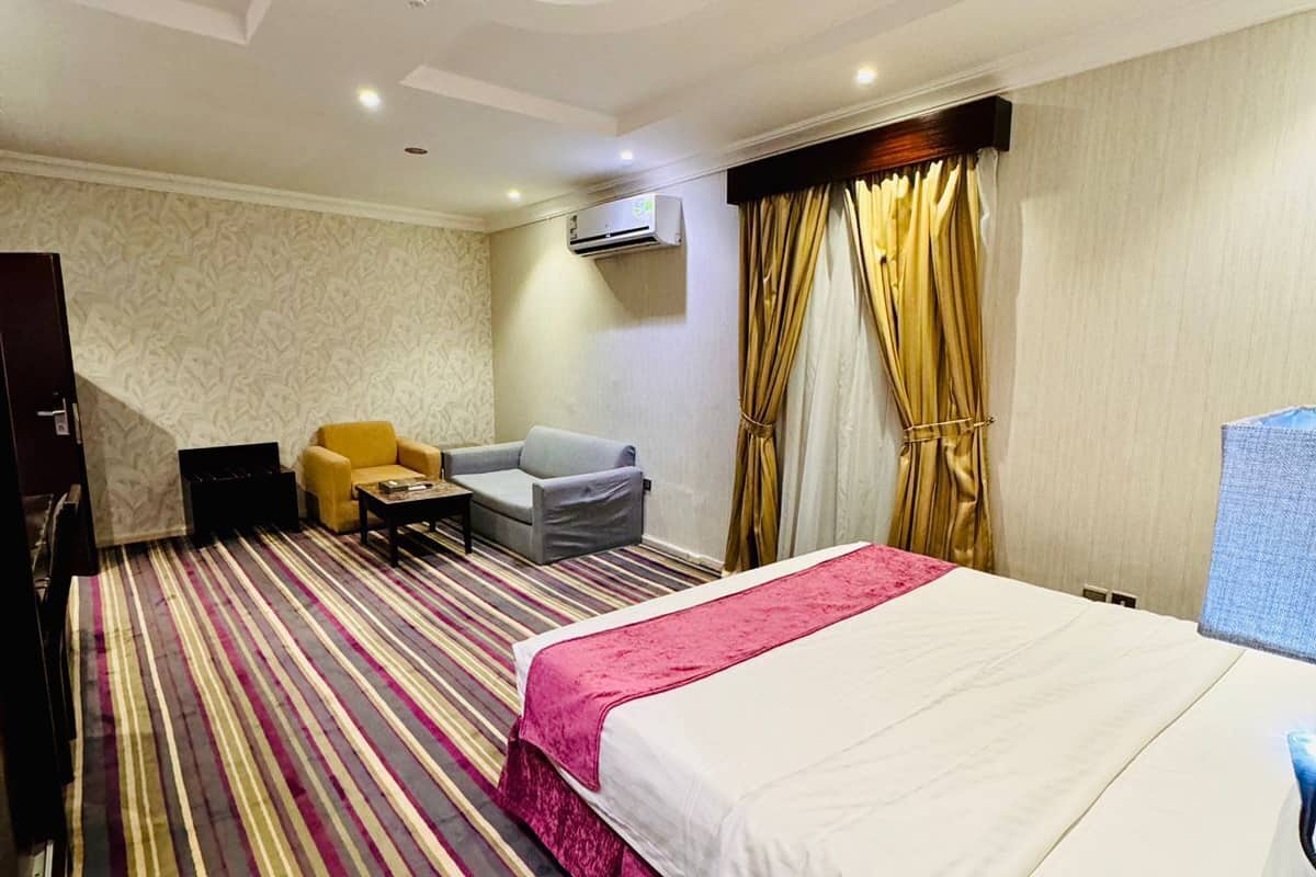 A hotel room with a bed, a desk with a telephone, a chair, a sofa, and an air conditioning unit.