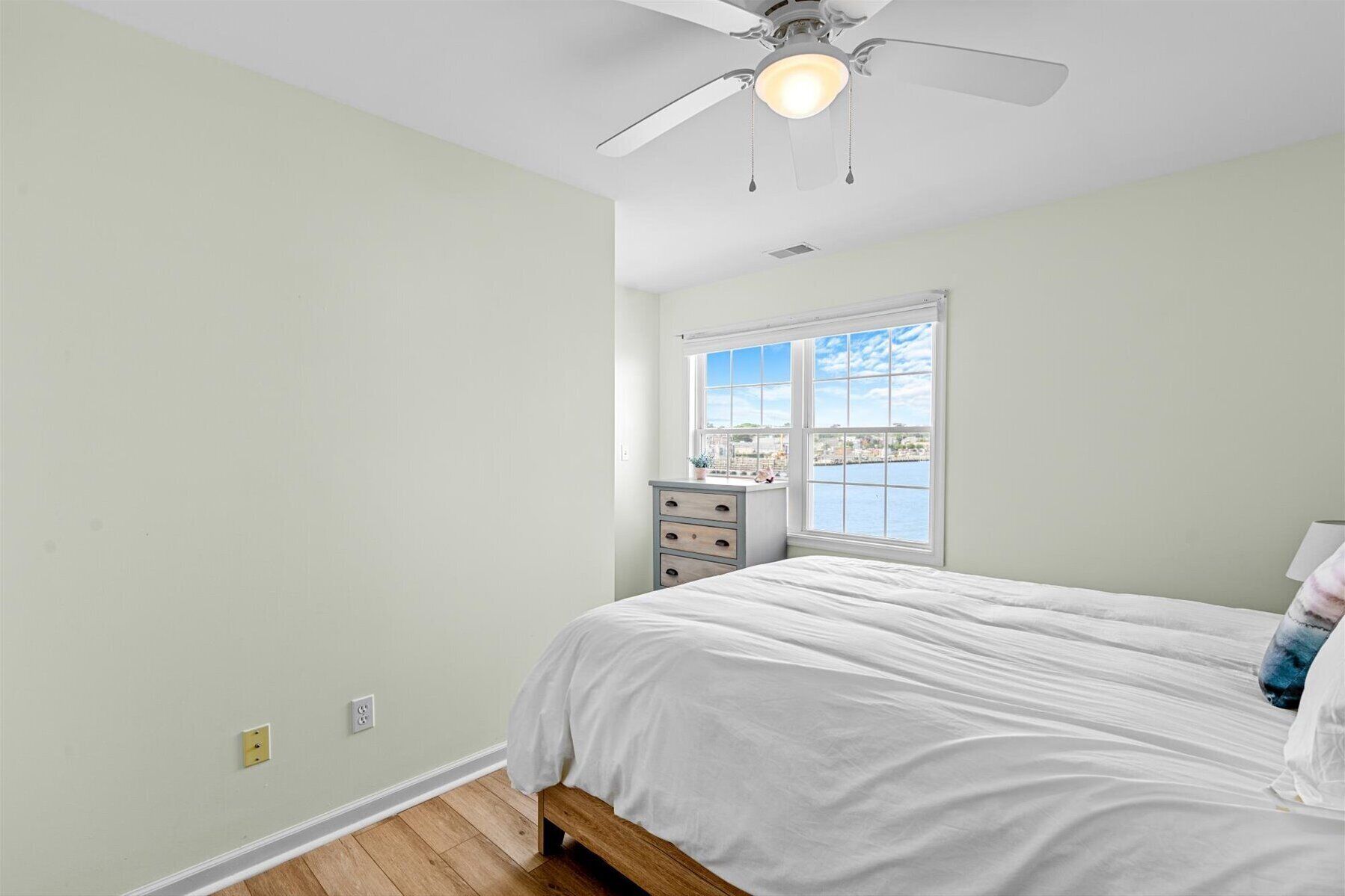 Quarto com cama, janela vista para a água, ventilador de teto e cômoda pequena.