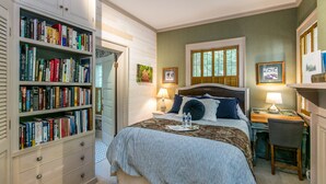 Un dormitorio con una amplia estantería, una cama con colcha a cuadros, un escritorio con silla y una ventana con contraventanas.
