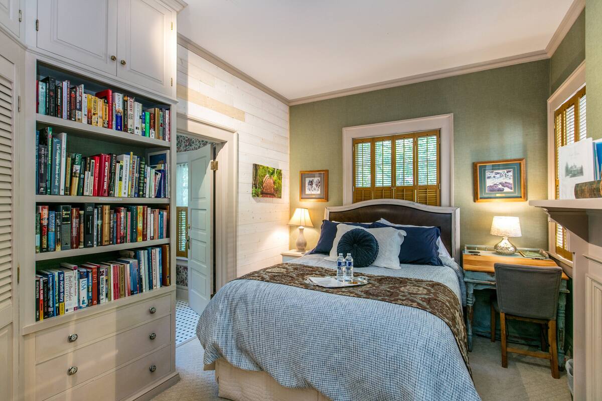 Een slaapkamer met een grote boekenkast, een bed met een geruit sprei, een bureau met stoel, en een raam met luiken.