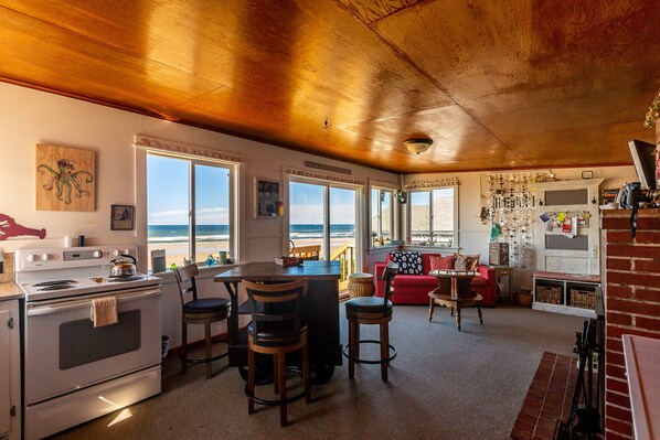 A kitchen with a stove, chairs, a dining table, and large windows overlooking the beach.