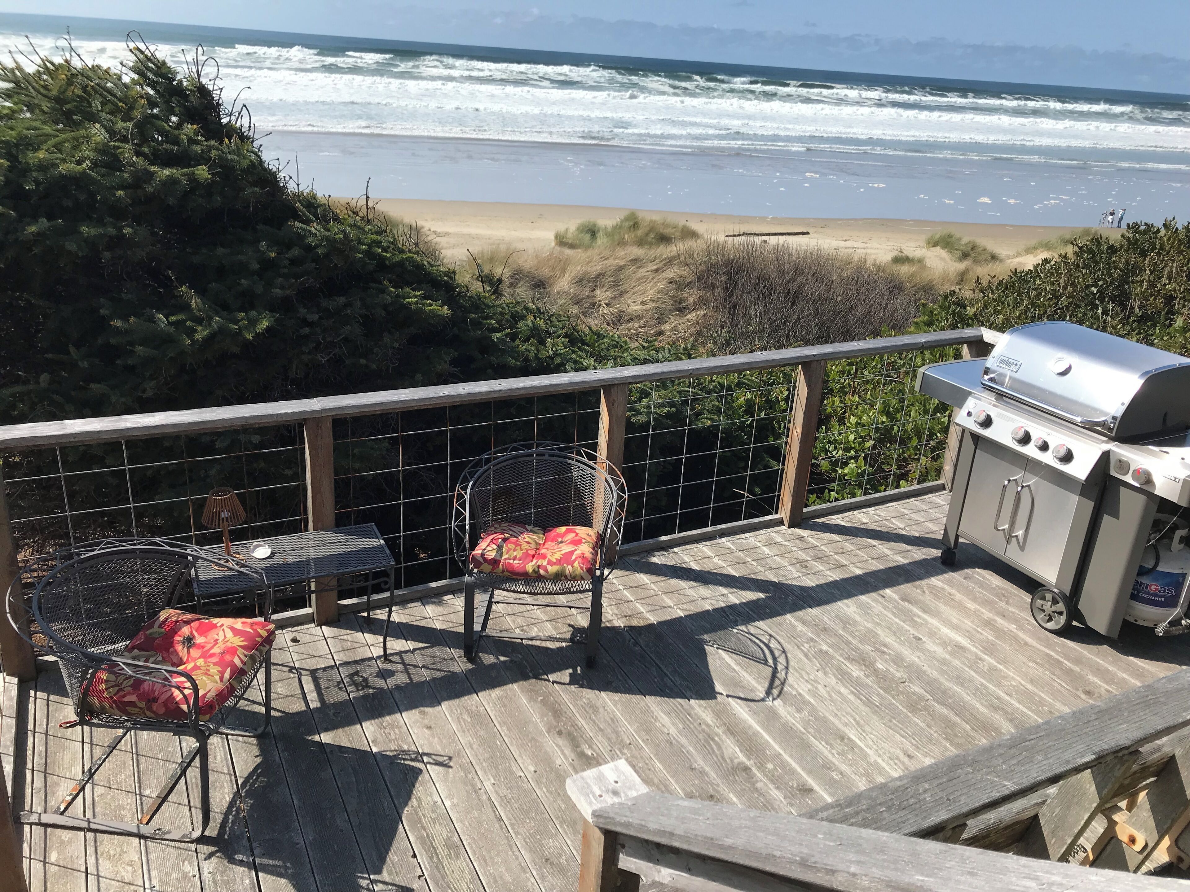 A deck with a grill, two wicker chairs, and a view of the beach.