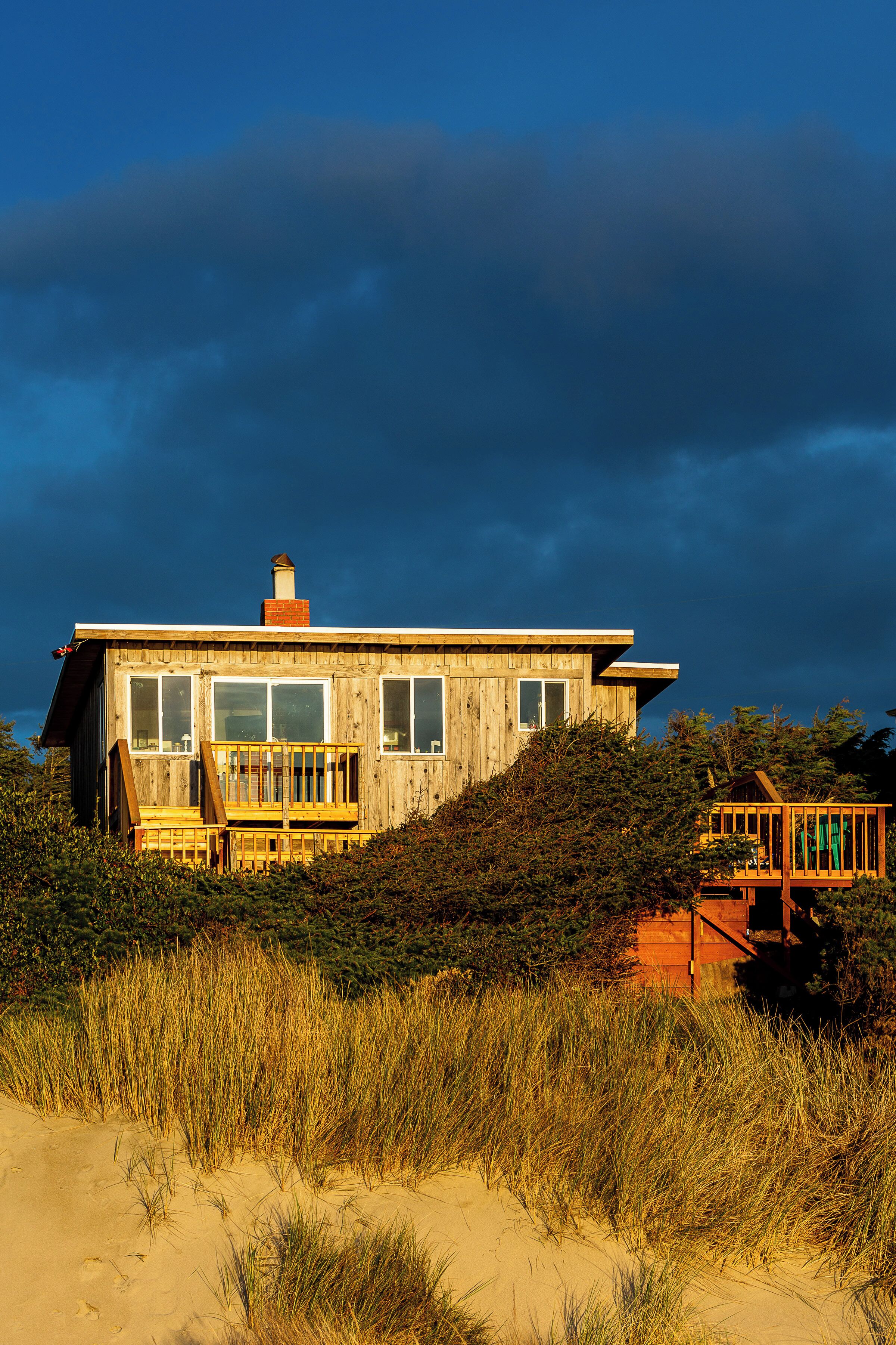 A wooden house with a deck, surrounded by sand and grass.