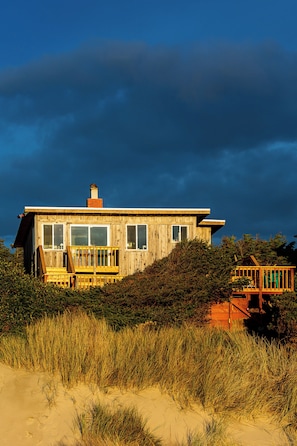 A wooden house with a deck, surrounded by sand and grass.