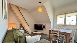 Une pièce de style « loft » avec un plafond en pente, un escalier en bois, un canapé vert, une table à manger avec des chaises et un téléviseur fixé au mur.