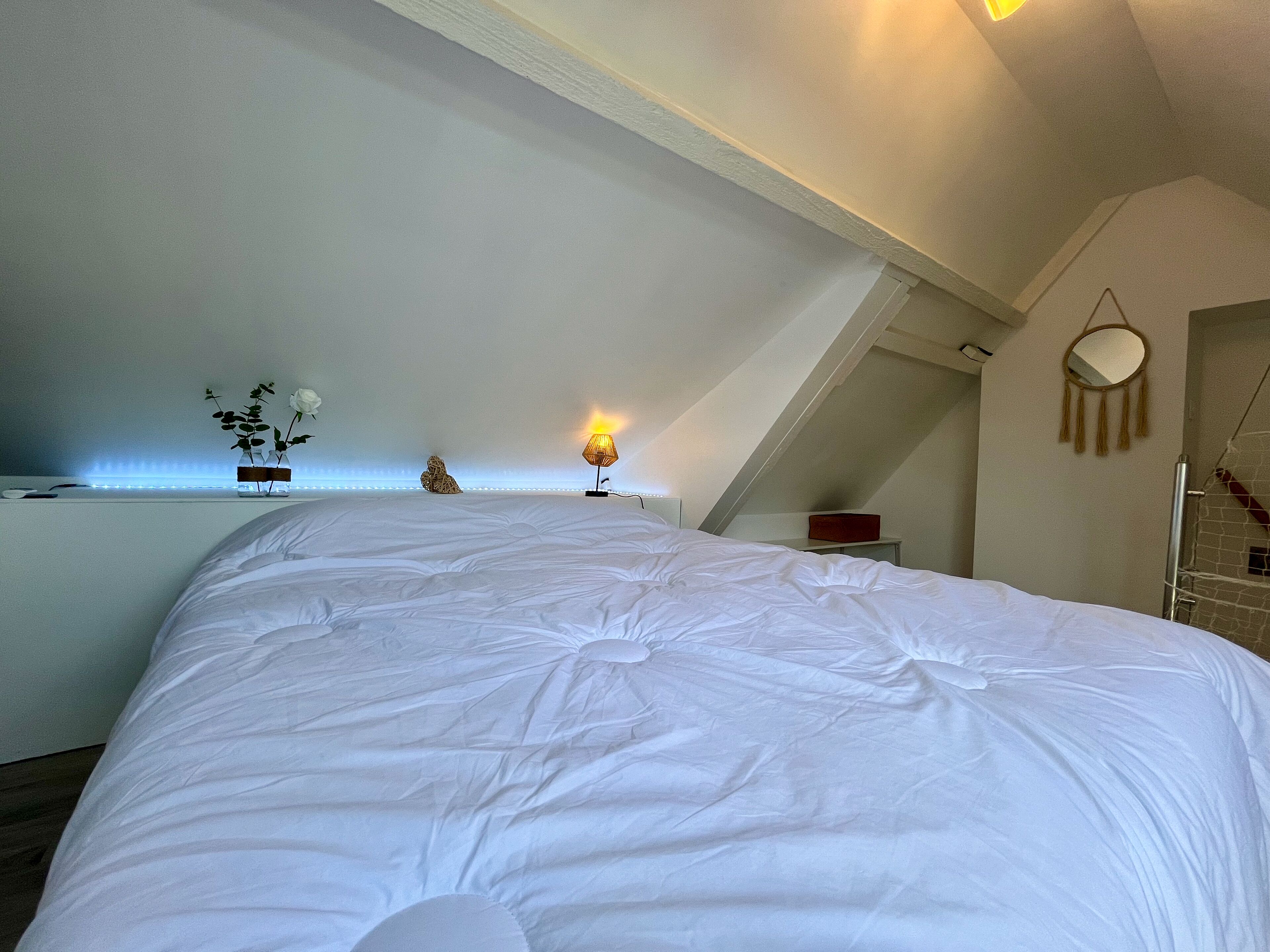 Een slaapkamer met een groot bed, een nachtkastje met een lamp, een spiegel aan de muur en een plank met decoratieve objecten.