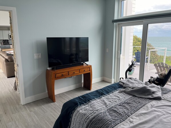 A bedroom with a large bed, a TV on a wooden stand, and a balcony with a view of the sea.