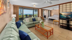 A spacious room with a large sofa, a coffee table, and a bed. There is a television and a ceiling fan. The room has large windows with a view of the ocean.